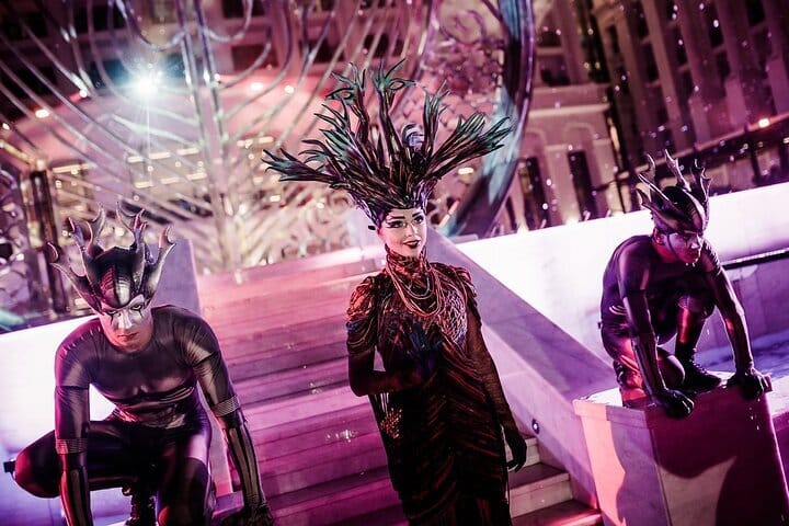 Three performers in dramatic and feathered headdesses and body-hugging costumes pose together.
