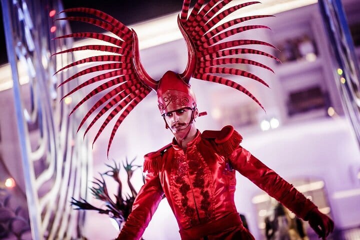 A man in a red military uniform with large feathrred headdress strikes a pose.